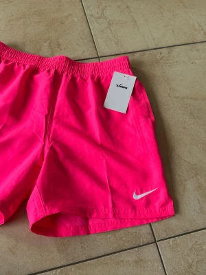 Neonrosa badshorts från Nike - Snygga neonrosa badshorts från Nike i storlek S med elastisk midja och vit Nike-logga på benet. Perfekta för stranden eller poolen, tillverkade i snabbtorkande material för maximal komfort. Klassisk modell med korta ben och enkel design. Säljer då det var fel storlek, de är alltså helt nya.