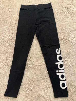 Svarta leggings från Adidas - Snygga svarta leggings från Adidas med stort vitt logotryck längs ena benet. De har en klassisk, tight passform och bred resår i midjan. Vet tyvärr ej vilket storlek men skulle gissa på XS 