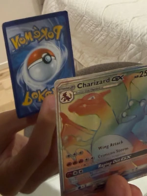 Charizard GX Pokémonkort - Säljer ett Charizard GX Pokémonkort med holografisk yta. Kortet har färgstark design med Charizard i orange, rött och blått. Text och symboler är tydliga på framsidan och baksidan har klassiskt Pokémon-motiv med Pokéboll. Plastficka kommer med kortet. Pris kan diskuteras