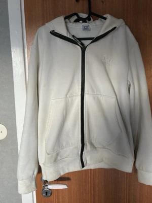 Vit hoodie från C.P. Company - Vit hoodie från C.P. Company med dragkedja framtill och klassiska goggles på huvan. Hoodien har två stora fickor fram och ribbade muddar vid ärmar och nederkant. Tillverkad i mjukt bomullsmaterial och har en broderad logga på bröstet.