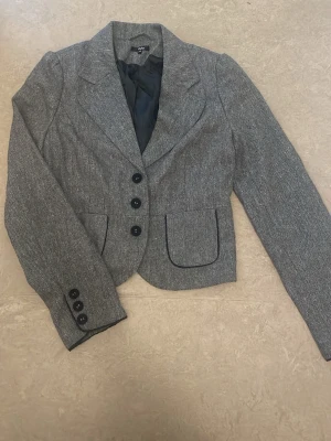 Grå kavaj med svarta knappar  - Snygg grå kavaj från cubus med fyra svarta knappar framtill och tre på ärmsluten. Kavajen har markerade fickor med svart kant och klassisk krage. Materialet är vävt i en diskret mönstrad struktur och helfodrad på insidan.