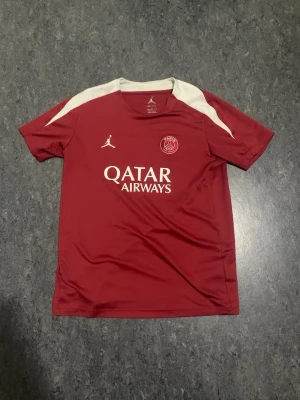 Röd PSG träningströja Jordan - Snygg röd PSG träningströja från Jordan med vita detaljer på axlarna. Tröjan har tryck med Qatar Airways och PSG-logga på bröstet samt Jordan-logga. Tillverkad i ett lätt och snabbtorkande material, perfekt för träning eller match.