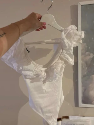 Vit body med volangärmar - Supersöt vit body med djup ringning och stora volangärmar. Tillverkad i mjukt, stretchigt tyg som sitter snyggt på kroppen. Perfekt för dig som vill ha något extra till outfiten. Ingen synlig logga eller märke.