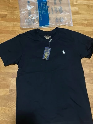 Svart t-shirt från Polo Ralph Lauren - Klassisk svart t-shirt från Polo Ralph Lauren med vit broderad logga på bröstet. T-shirten har korta ärmar och v-ringad hals. Tillverkad i mjuk bomull för en bekväm känsla och stilren look.