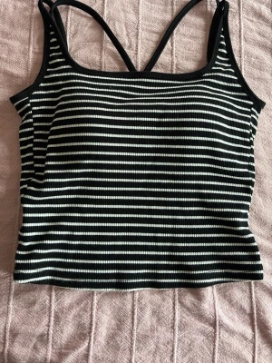 Randig ribbad linnetopp med vaddering  - Säljer en svartvit randig linnetopp med smala axelband som korsas i ryggen. Toppen är croppad och ribbad vilket ger en snygg och trendig look. Perfekt till jeans eller kjol för en chill stil.