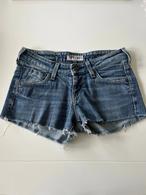 Blå jeansshorts från Lee - Säljer ett par klassiska blå jeansshorts från Lee med råa, fransiga kanter och femficksmodell. Shortsen har låg midja och är perfekta för varma sommardagar! 