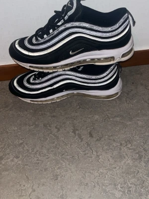 Nike Air Max 97 svart/vit sneakers - Nike Air Max 97 sneakers i svart med vita och silvriga vågformade detaljer. Skorna har en synlig Air Max-sula och reflekterande partier längs ovandelen. Klassisk låg modell med snörning och ikonisk design som sticker ut.