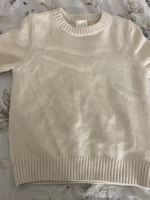 Krämvit stickad tröja från H&M - En enkel och stilren krämvit stickad tröja från H&M.  Tröjan har rund halsringning, ribbade muddar vid ärmslut och nederkant samt en mjuk och bekväm känsla. Perfekt basplagg för lager på lager och passar till det mesta.