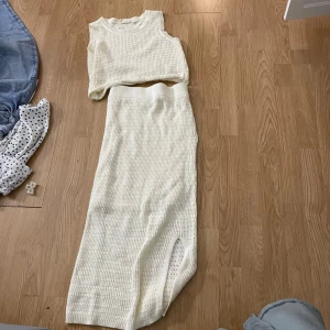 Stickat vitt set med topp och kjol - Säljer ett snyggt stickat set i vitt med ärmlös topp och lång kjol. Perfekt till stranden eller vardagligt. Setet har ett hålmönstrat material och bred resår i midjan på kjolen. Perfekt för dig som gillar en avslappnad men trendig stil.