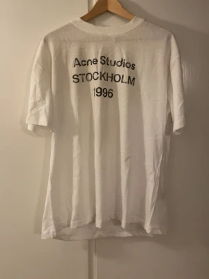 Vit Acne Studios t-shirt Stockholm - Vit t-shirt från Acne Studios med svart text 'Acne Studios STOCKHOLM 1996' på bröstet. Klassisk rak passform och rund halsringning. Tillverkad i mjuk bomull, perfekt för en clean och stilren look.