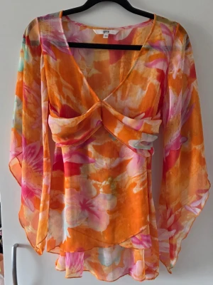 Blommig transparent blus - Färgstark blus i transparent material med stora blommor i orange, rosa och vitt. Djup V-ringning, volangdetaljer framtill och knytband i ryggen. Långa, flödande ärmar och luftig passform ger en bohemisk vibe. Perfekt för dig som gillar statement-plagg.