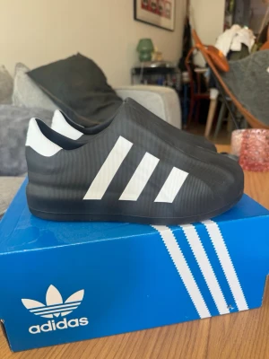 Svarta Adidas tofflor med vita ränder - Säljer ett par svarta Adidas tofflor med de klassiska tre vita ränderna på sidan. Tofflorna har en modern, chunky design och är tillverkade i ett gummiliknande material som gör dem både lätta och bekväma att bära. Perfekta för chill dagar hemma eller snabba ärenden.