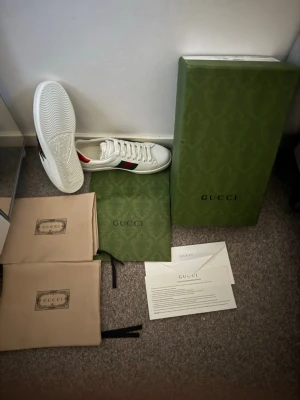 Gucci Ace sneakers med orm - Säljer ett par vita Gucci Ace sneakers i läder med den ikoniska gröna och röda webbranden samt orm-motiv på sidan. Skorna har metallic-detaljer i rött och grönt på hälen och klassisk låg siluett med vita snören. Kommer med originalkartong och dustbags. Helt nya kvitto finns!!🙌🙌