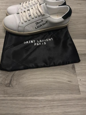 Saint Laurent vita sneakers -  Säljer ett par vita Saint Laurent sneakers i läder med svart detalj på hälen och handskriven logga på sidan. Klassisk låg modell med vita snören och gummisula. Kommer med original dustbag. Använda ca 5 gånger. Väldigt gott skick!