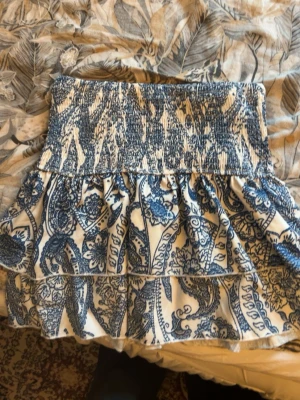 Blåvit kortkjol med volanger - Supersöt kortkjol i blått och vitt med smock i midjan och volangdetaljer. Kjolen har ett unikt mönster och är perfekt för dig som vill ha en lätt och luftig stil. Passar perfekt till sommaren och ger en fräsch vibe till din outfit.