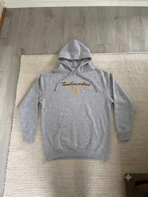 Grå Timberwolves hoodie XL - Snygg grå hoodie från Timberwolves med broderad gul text och logga på bröstet. Klassisk känguruficka framtill och justerbar huva med dragsko. Tillverkad i mjuk bomullsblandning (80% bomull, 20% polyester) för skön känsla.