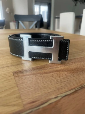 svart läderbälte H-spänne - Snyggt svart bälte i äkta läder med kontrastsömmar och ett stort, silverfärgat H-spänne i metall. Perfekt accessoar för att lyfta vilken outfit som helst med en exklusiv touch.