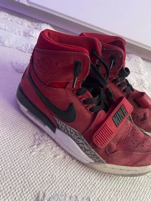 Röda Nike Air Jordan sneakers - Säljer ett par röda Nike Air Jordan 312 torro sneakers med svarta detaljer och klassisk Nike-logga på sidan. Skorna har högt skaft, svart snörning och kardborreband med Nike-tryck. Mellansulan har ett grått mönster och ovandelen är i läder. Perfekta för dig som gillar streetwear. Tyvärr så finns det lite märken på det svarta vid sulan men det är bara att måla över med svart färg. Inget som skadar skorna! 