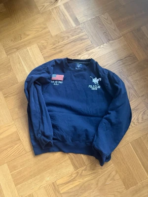 Mörkblå sweatshirt Polo Club Vinson - Mörkblå sweatshirt från Polo Club Vinson med broderad logga och polospelare på bröstet. Tröjan har även en amerikansk flagga och texten 'San Antonio U.S.A.' på ena sidan. Klassisk rund hals och ribbade muddar. Perfekt för en avslappnad stil.