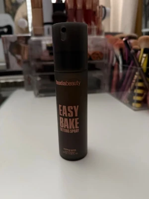 Huda Beauty Easy Bake Setting Spray - Face mist från Huda Beauty i en matt svart flaska med rosa text. Produkten heter Easy Bake Setting Spray och innehåller risstärkelse, niacinamid och hyaluronsyra. Alkoholfri och parfymfri formula som ger en fräsch finish. Perfekt för att fixera makeup hela dagen.