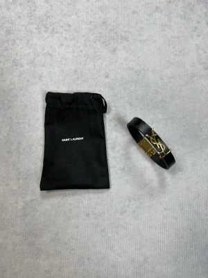 Svart läderarmband från Saint Laurent - Snyggt svart armband i läder från Saint Laurent med ikoniskt YSL-monogram i guldfärgad metall. Armbandet har en stilren och minimalistisk design som passar perfekt till din outfit. Levereras med originalpåse.