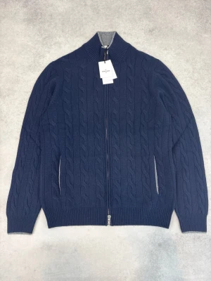 Marinblå kabelstickad ziptröja Gran Sasso - Kabelstickad marinblå tröja från Gran Sasso med hel dragkedja framtill och ribbstickad grå krage. Tröjan har långa ärmar, ribbade muddar och två sidofickor. Perfekt för lager på lager och snygg till jeans eller chinos.