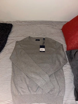 Grå stickad tröja från GANT - Snygg grå stickad tröja från GANT med rund halsringning och diskret logga broderad på bröstet. Tröjan har långa ärmar och ribbade muddar vid ärmslut och nederkant. Perfekt för lager-på-lager och passar till det mesta.  Storlek S 