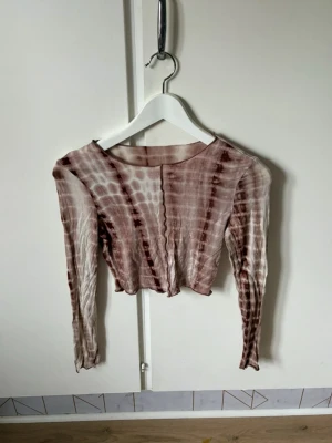 Brun tie-dye croppad långärmad topp - Trendig croppad långärmad topp i brun och beige tie-dye. Tunn och stretchig med vågig kant nertill och längs ärmarna. Perfekt för dig som gillar unika mönster och en avslappnad stil.