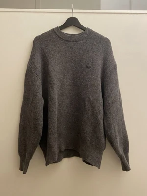Grå Lacoste Stickad Tröja (Nyskick) - Säljer en riktigt snygg och stilren stickad tröja från Lacoste i en mörkgrå melerad färg. Tröjan har den klassiska krokodilloggan broderad på bröstet i en diskret matchande färg (ton-i-ton), vilket ger ett exklusivt intryck. Tröjan är oanvänd utan prislapp, 10/10 skick.