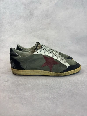 Golden Goose sneakers med röd stjärna - Snygga sneakers från Golden Goose i grått mocka med vit läderdetalj, svart häl och tå samt en ikonisk röd stjärna på sidan. Skorna har vita snören och en gulnad sula för en vintage look. Perfekta för dig som gillar streetstyle och unika detaljer.