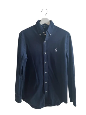 Mörkblå skjorta från Ralph Lauren - Klassisk mörkblå skjorta från Ralph Lauren med button-down krage och vita knappar. Skjortan har lång ärm och den ikoniska vita broderade loggan på bröstet. Perfekt för en stilren look och tillverkad i mjukt bomullstyg.