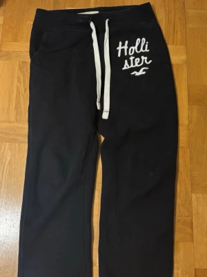 Svarta mjukisbyxor från Hollister - Mjukisbyxor från Hollister med vita dragsnören och broderad logga på vänster sida. Byxorna har avslappnad passform, resår i midjan och mjukt material. *OBS! Jag har klippt byxorna längst nere så att dom skulle passa bättre i längden på mig som är 157cm! 