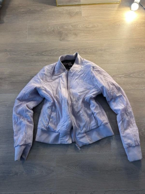 Ljuslila bomberjacka från Lululemon - Snygg Lululemon ljuslila bomberjacka med klassisk ribbad krage, muddar och midja. Jackan har två sidofickor och stängs med dragkedja framtill. Materialet är glansigt och ger en modern look. Perfekt för dig som vill sticka ut med en unik färg.