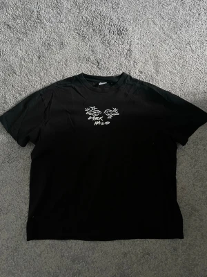 Svart Dark Halo t-shirt med ängel - Cool svart t-shirt från Dark Halo med vit grafisk text och motiv framtill samt en stor airbrush-inspirerad ängel på ryggen. Oversized passform och tillverkad i mjuk bomull. Perfekt för dig som gillar streetwear och unika prints.