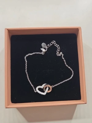 Edblad armband med hjärta och ring - Snyggt armband från Edblad i silverfärgat metall med ett gnistrande hjärta dekorerat med små stenar och en slät ring i roséguldston. Tunn kedja och justerbar längd. Perfekt accessoar för dig som gillar stilrena smycken med en romantisk touch.