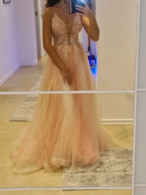 Lång rosa balinspirerad klänning - Romantisk långklänning i ljusrosa med spetsdetaljer och djup v-ringning. Kjolen är i flera lager tyll för en fluffig look och livet har broderade blommor. Axelband design med markerad midja, perfekt för speciella tillfällen.Bal eller bröllopsgäst.