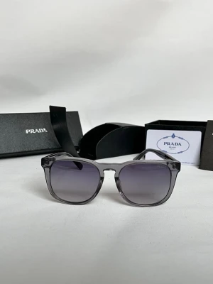 Gråa solglasögon från Prada - Snygga solglasögon från Prada med transparent grå båge och mörktonade glas. Klassisk fyrkantig form och röda detaljer med Prada-logga på skalmen. Perfekt accessoar för dig som vill ha en stilren och modern look.