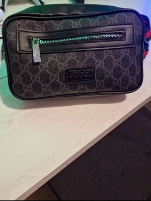 Gucci axelväska med monogram - Snygg axelväska från Gucci i svart och grått med klassiskt GG-monogram. Väskan har en framficka med silverfärgad dragkedja och en bred axelrem i rött och marinblått. Tillverkad i canvas med detaljer i läder. Perfekt för dig som gillar streetstyle och designeraccessoarer. 1:1