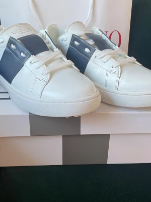 Vita sneakers, Valentino, strl 42 - Snygga vita sneakers från Valentino Garavani med bred mörkblå läderdetalj över sidan och nitar på hälen. Klassisk låg modell med vita skosnören och diskret logga på plösen. Perfekta för dig som gillar stilrena och exklusiva sneakers. 