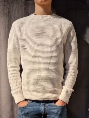 Vit ribbstickad tröja - Säljer en stilren vit ribbstickad tröja med rund halsringning och långa ärmar. Tröjan har en enkel och modern design med breda muddar vid ärmslut och nederkant. Perfekt för lager-på-lager och passar till jeans eller chinos.