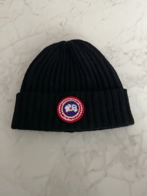 Svart ribbstickad mössa Canada Goose - Svart ribbstickad mössa från Canada Goose med klassisk uppvikt kant och det ikoniska runda Arctic Program-märket framtill. Märket är broderat i rött, vitt och blått. Perfekt för kalla dagar och ger en clean streetwear-look.