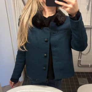 Mörkblå kappa med pälskrage - Snygg mörkblå kappa med svart fuskpälskrage och stora knappar framtill. Perfekt för dig som vill ha en klassisk men trendig look. Köpt på Sellpy, ej använd. Lik stilar som Meotine, Zara, Arket osv
