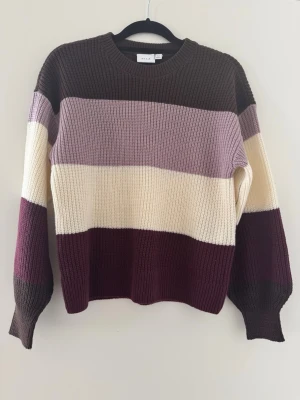 Randig stickad tröja från Vila - Stickad tröja från Vila med breda horisontella ränder i brunt, lila, cremevitt och vinrött. Tröjan har rund halsringning, långa ärmar och ribbstickade muddar. Perfekt för lager-på-lager och en färgglad look.