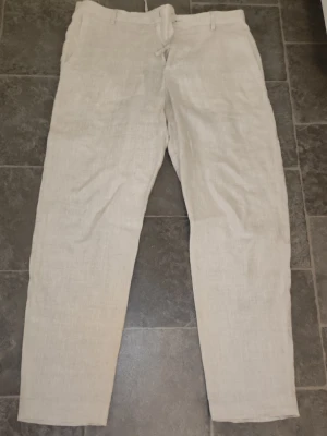 Beiga linnebyxor från Zara - Snygga beiga linnebyxor från Zara med rak passform och dragsko i midjan. Byxorna har bälteshällor och elastisk midja baktill för extra komfort. Perfekta för en avslappnad och stilren look under varmare dagar.