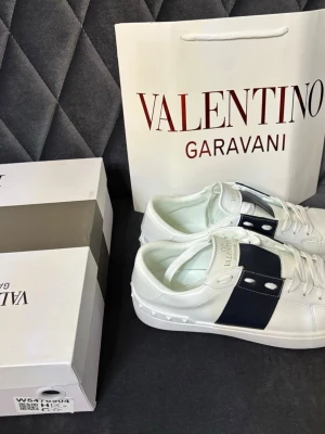 Valentino Garavani vita sneakers - Stilrena vita sneakers från Valentino Garavani med bred svart läderdetalj och nitar på sulan. Klassisk låg modell med vita skosnören och guldfärgad logga på plösen. Perfekta för dig som gillar lyxig och modern streetstyle.