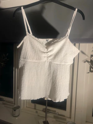 Vitt linnetopp med tunna band - Säljer en söt vit linnetopp med smockat tyg och tunna axelband. Toppen har en lätt rynkad detalj framtill och vågig kant nertill. Perfekt till sommaren och enkel att matcha med jeans eller kjol.💕 tveka inte att fråga om du har några frågor😊