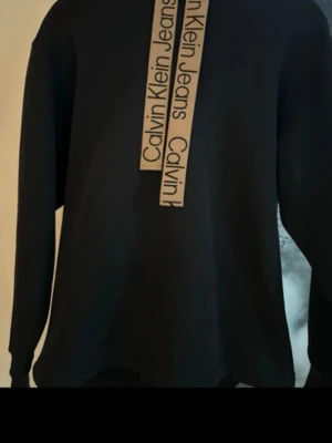 Svart hoodie från Calvin Klein Jeans - Snygg svart hoodie från Calvin Klein Jeans med två beige band framtill där loggan står tryckt i svart. Hoodien har en klassisk passform och långa ärmar. Perfekt för en avslappnad och trendig look.