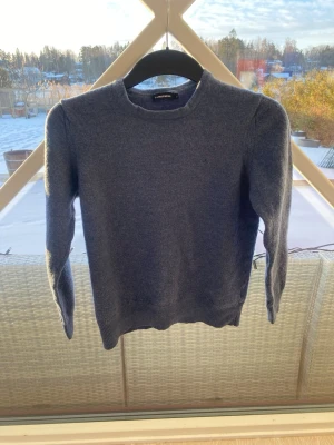 Mörkgrå stickad tröja J.Lindeberg - Mysig mörkgrå stickad tröja från J.Lindeberg med rund halsringning och diskret broderad logga på bröstet. Tröjan har långa ärmar och ribbade muddar vid ärmslut och nederkant. Perfekt för kyliga dagar och enkel att matcha.