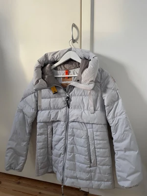 Ljusgrå dunjacka från Parajumpers - Unik ljusgrå/blå dunjacka från Parajumpers med hög krage, dragkedja och två stora fickor framtill. Jackan har en ikonisk gul detalj vid halsen och ett broderat märke på ärmen. Sista bilderna visar lite defekter av smink men det syns inte när man har den på sig. 