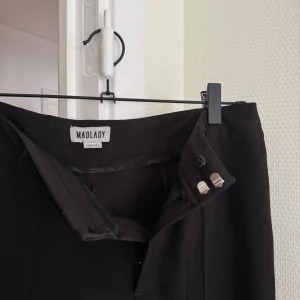 Svarta kostymbyxor från Madlady - Svarta kostymbyxor från Madlady med raka ben och hög midja. Byxorna har pressveck framtill, bälteshällor och stängs med dragkedja och två knappar. 